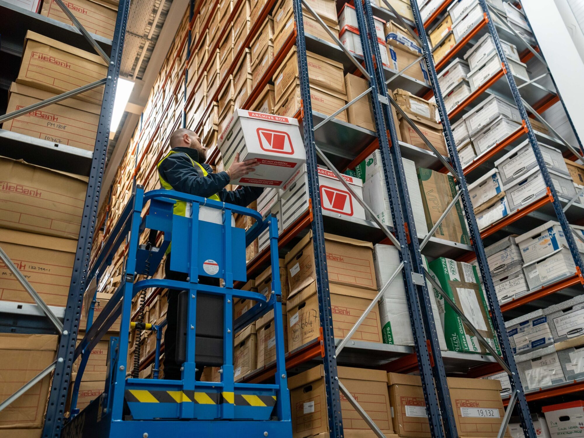 A photo of a Vanguard employee standing on a mobile lift placing a Vanguard cardboard box on a shelf.