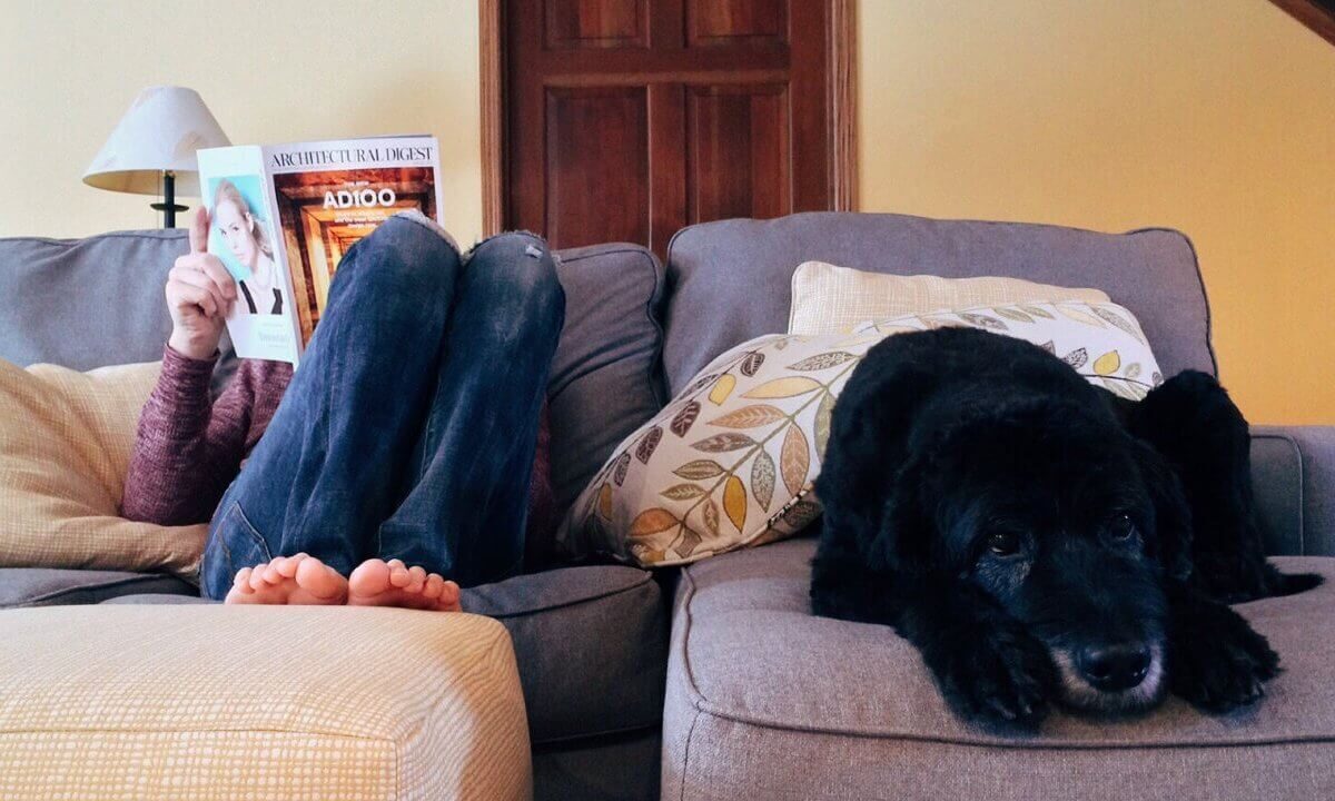 A photo showing a woman reading a magazine on the sofa with a dog laying next to her.