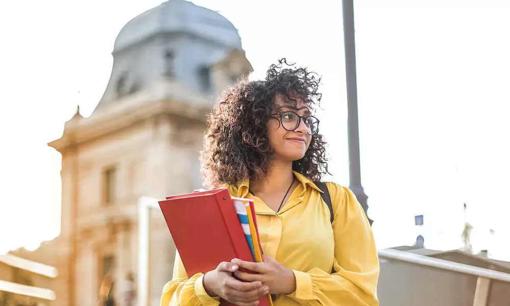 A photo of a student waring a backpack and holding a binder and notebooks.