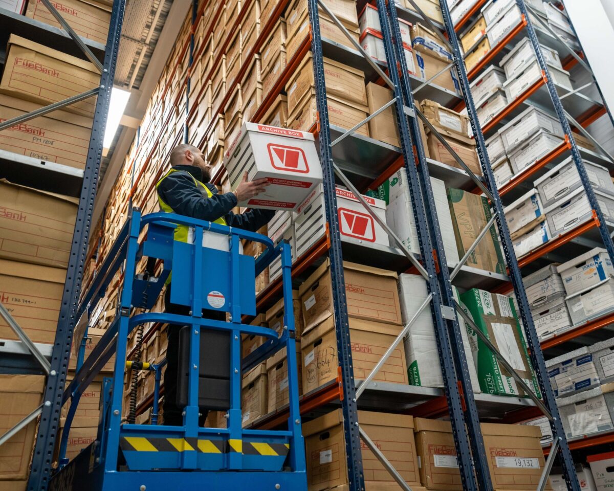 A photo of a Vanguard employee standing on a mobile lift placing a Vanguard cardboard box on a shelf.
