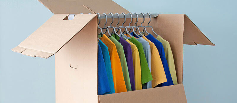 A photo showing a cardboard box specifically designed for the transport of garments on hangers. The photo shows some colourful t-shirts hanging inside of the box.