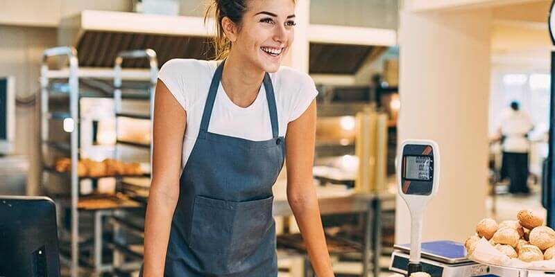 A photo showing a woman wearing an apron smiling and standing inside of a bakery, with some bread rolls and a scale next to her.