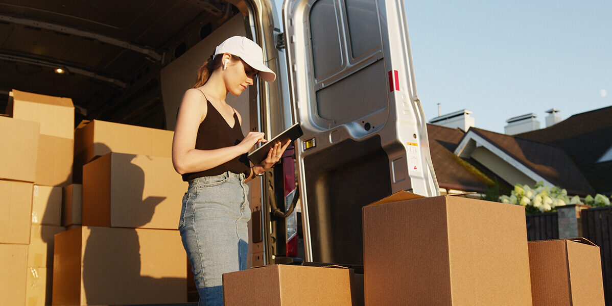 A photo showing a woman holding a pen and paper while loading up many cardboard boxes on a van.