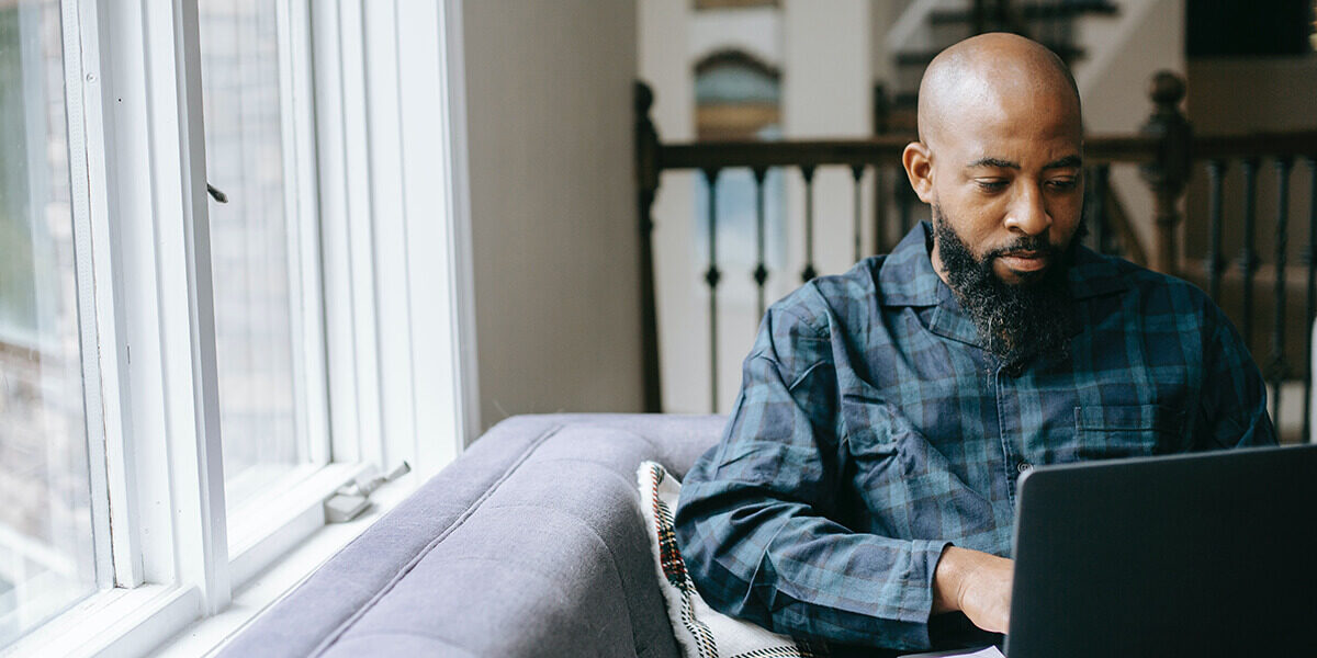 A photo of a man sitting on a chair by a window and working on his laptop.