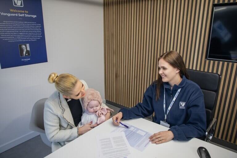 A photo showing a woman with a child and a Vanguard employee filling in paperwork in the reception of one of the Vanguard self storage branches.