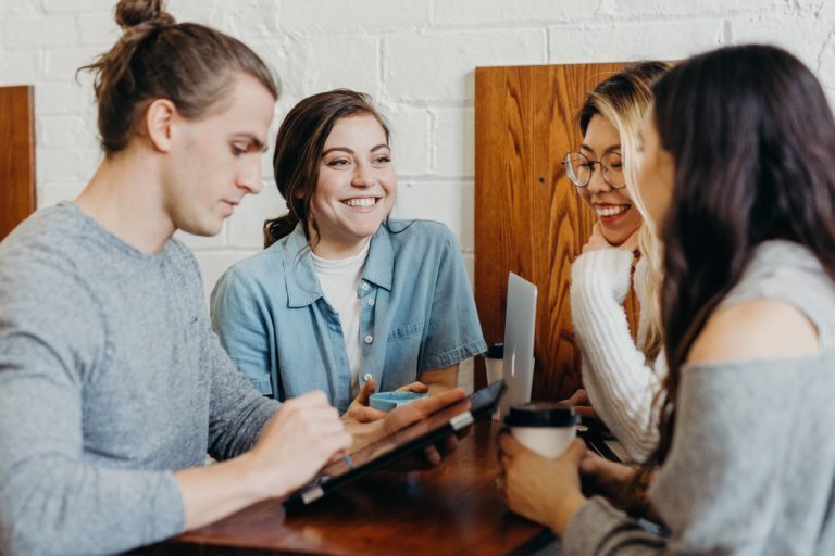 A photo showing a group of four people chatting with coffees in their hands. One of the four is holding a tablet in his hand and looking at it.
