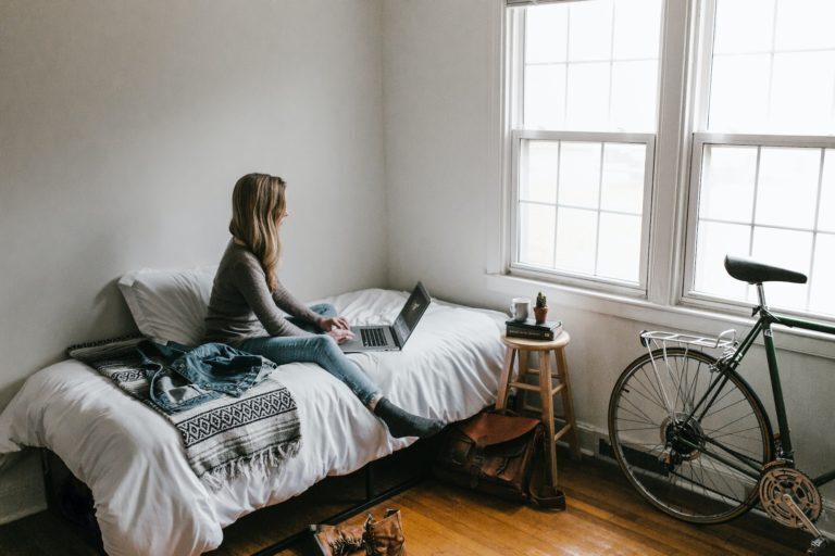 A photo showing a woman using her laptop on her bed, with a bedside table and a bike next to two large windows.